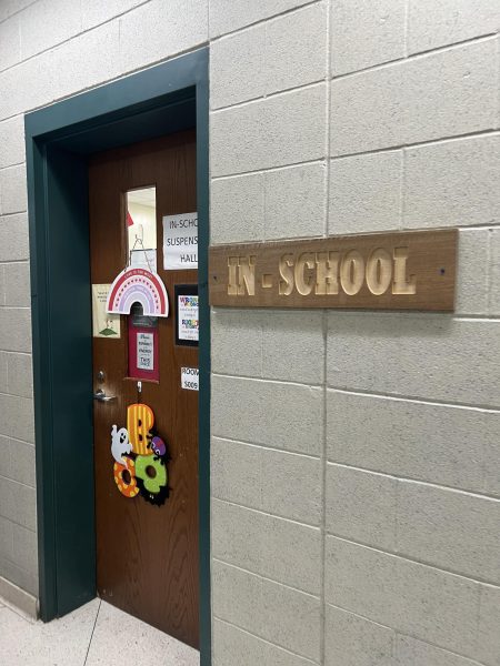 A photograph of the outside of a room with a sign made of wood. The sign has the words "In-School" carved into them. On the door there are a variety of different decorations, including a piece of white printer paper with the text "In-School Suspension Hall" written in all caps with black text.