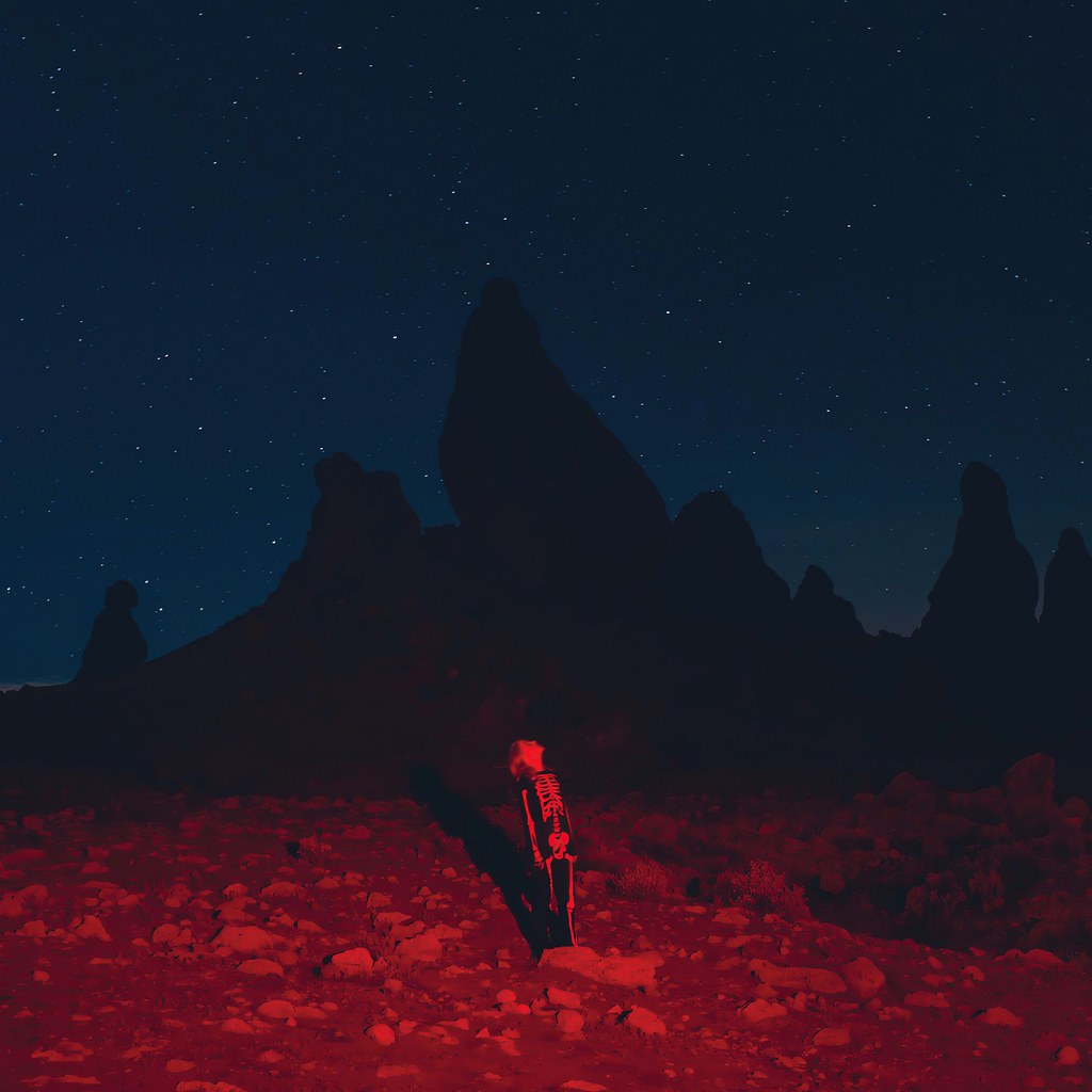 Phoebe Bridgers is standing in the middle of a field. In the background, there is a shadow of some mountains with a starry night sky behind it. Phoebe and the ground around her is surrounded by red light.